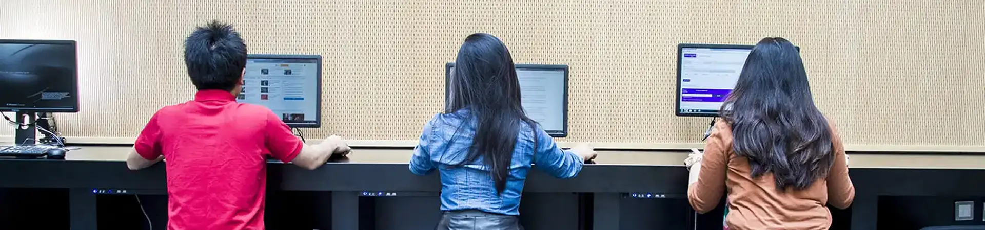 Three students working on desktop computers in a modern workspace, seen from behind.