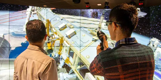 Two students in front of a large screen showing a spacecraft over earth