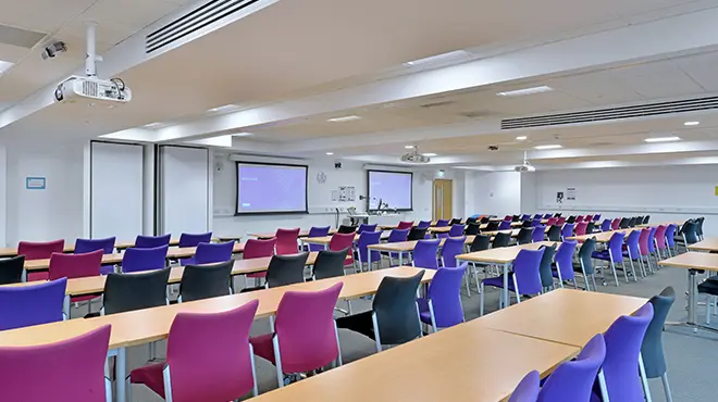 Classroom 3.006a/b at Alliance Manchester Business School, set up in a lecture-style layout with rows of tables and colourful chairs, presentation screens, and ceiling-mounted projectors.