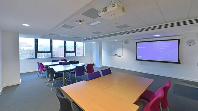 Classroom 3.008 at Alliance Manchester Business School, arranged in a boardroom-style layout with tables and chairs, a presentation screen, ceiling-mounted projector, and windows providing natural light.