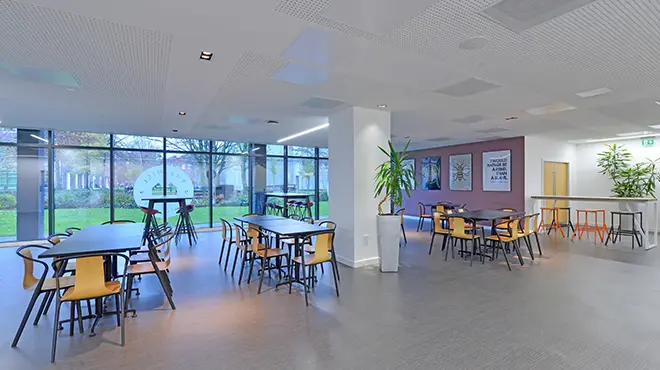 Interior of The Mill restaurant at Alliance Manchester Business School, showing a modern dining area with tables and chairs, indoor plants, and large windows overlooking a green courtyard.