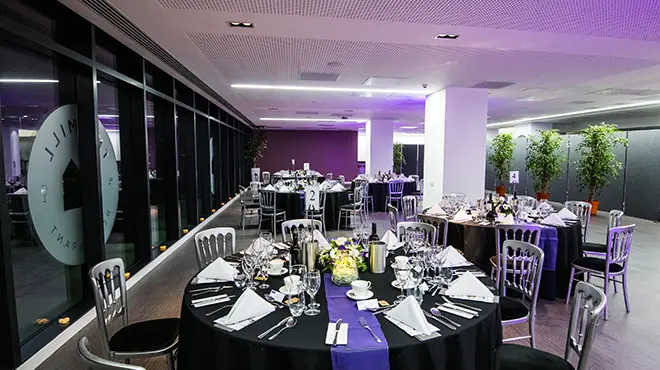 Interior of The Mill at Alliance Manchester Business School set up for a formal dinner, with round tables dressed in black linens, place settings and glassware, silver chairs, floral centrepieces, and ambient purple lighting.