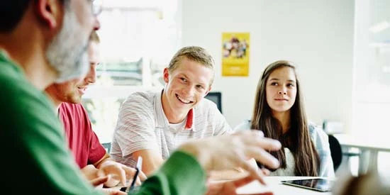 Smiling students sitting at table in high school classroom listening to an adult talking