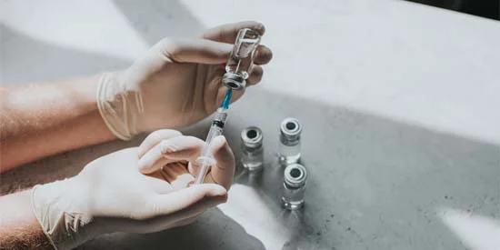 A hand wearing latex gloves holding a phial of clear liquid and a syringe