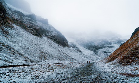 A region of mountains covered in snow with two people in the distance on the path at the foot of a mountain