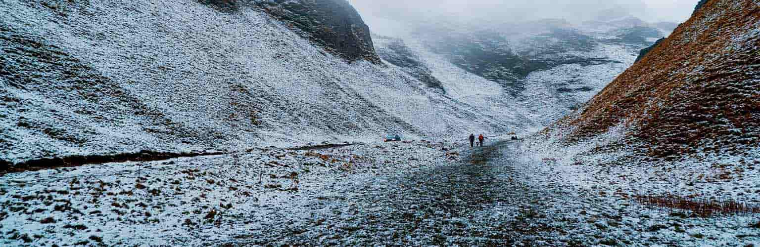 A region of mountains covered in snow with two people in the distance on the path at the foot of a mountain