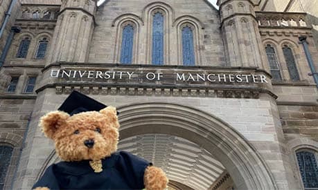 A teddy bear in a graduation cap and gown in front of the university of Manchester main building