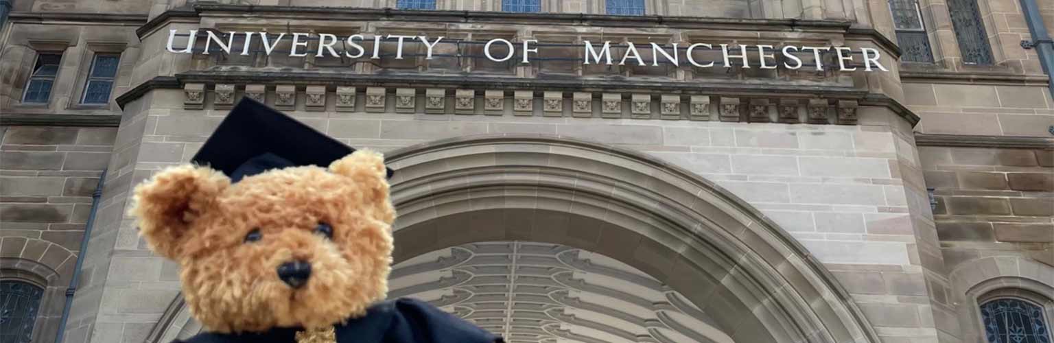 A teddy bear in a graduation cap and gown in front of the university of Manchester main building