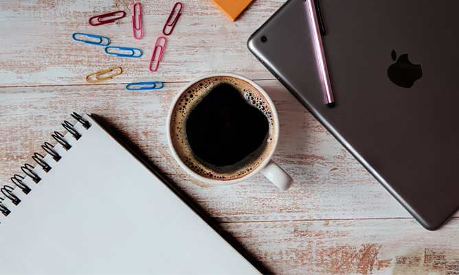 A wooden desk with a cup of coffee, a notepad and paperclips on.