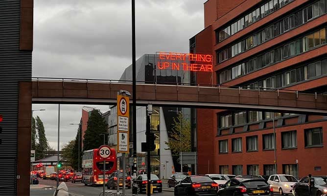 A view looking up Booth Street East in Manchester with a neon sign saying 'everything up in the air'