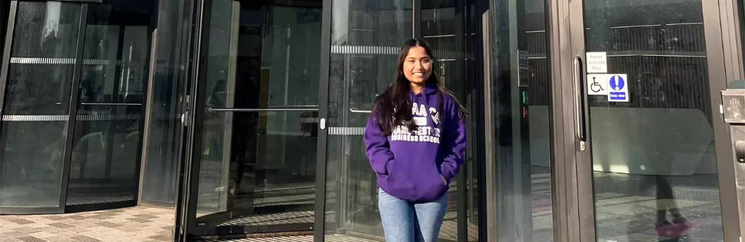 Hardika Gupta standing outside the entrance of the Alliance Manchester Business School building entrance
