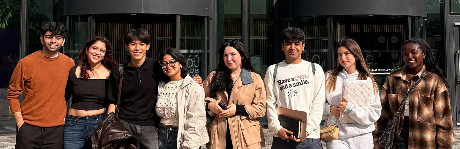 Group of eight young adults standing side by side outdoors in front of the Alliance Manchester Business School building, smiling and casually dressed, some holding notebooks or bags.