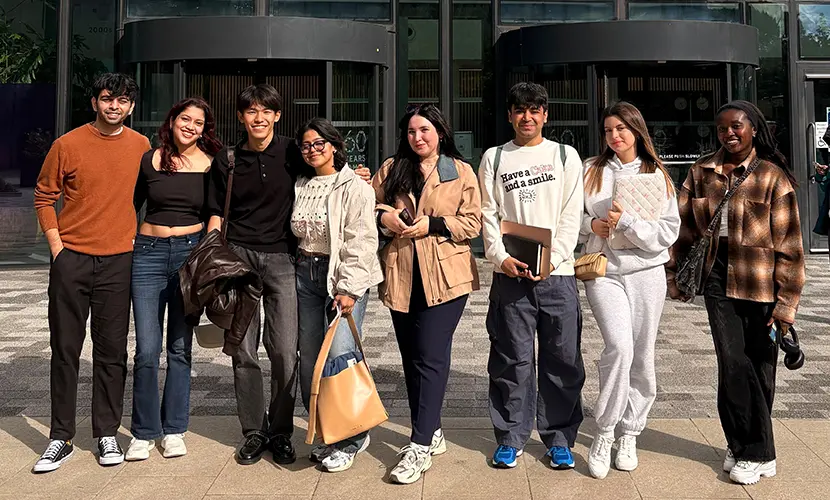 Group of eight young adults standing side by side outdoors in front of the Alliance Manchester Business School building, smiling and casually dressed, some holding notebooks or bags.