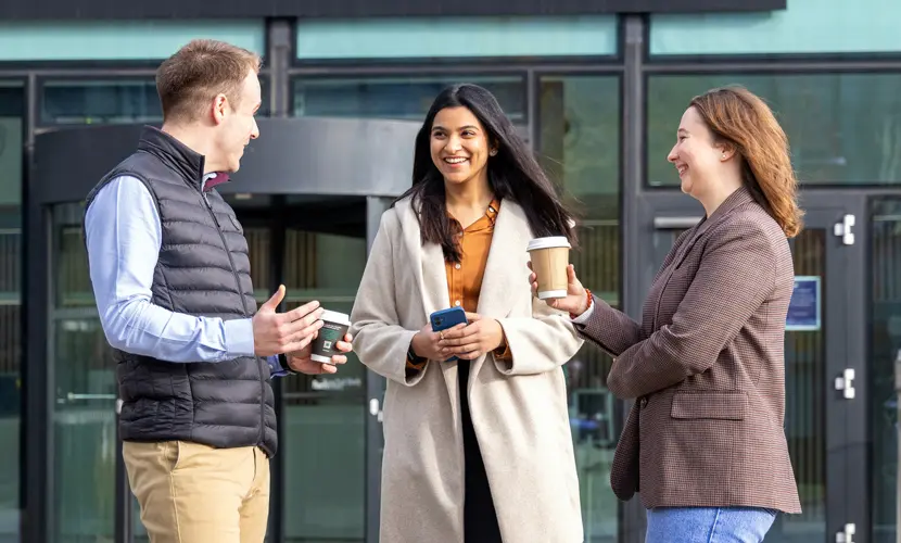 Three students outside the AMBS building chatting and drinking coffee