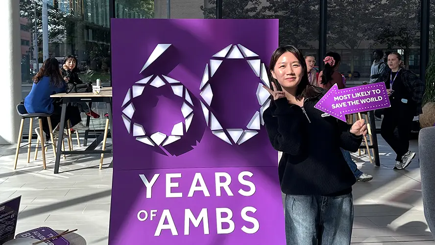 Pinyu Chen smiling next to a large purple sign that says 60 years of AMBS.