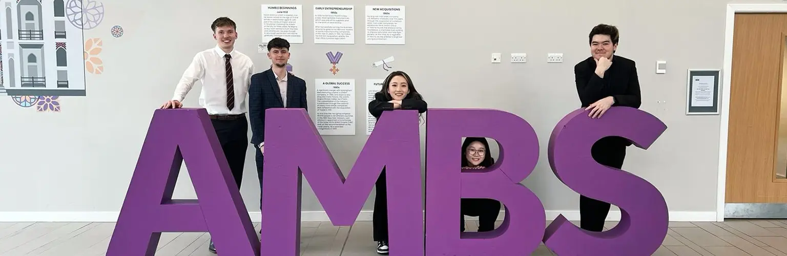 Five undergraduate students smiling for a photo between large letters that spell out AMBS.