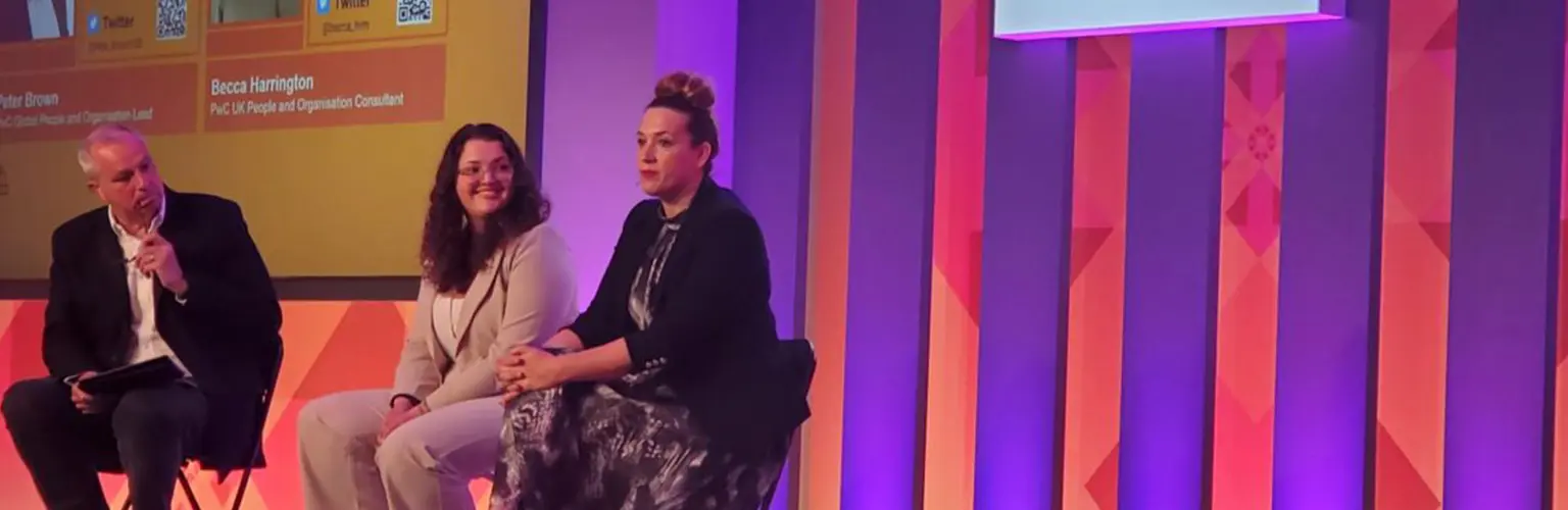 Panel discussion at a PwC event with three speakers seated on stage, PwC logo displayed on a large screen behind them.
