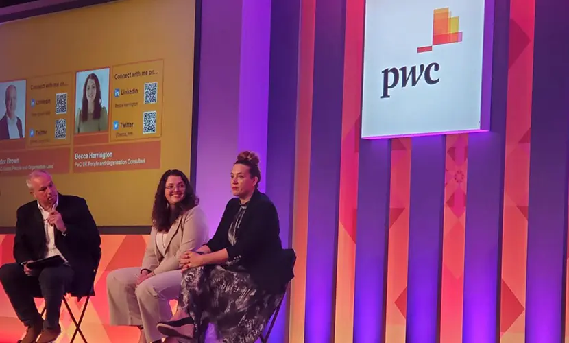 Panel discussion at a PwC event with three speakers seated on stage, PwC logo displayed on a large screen behind them.