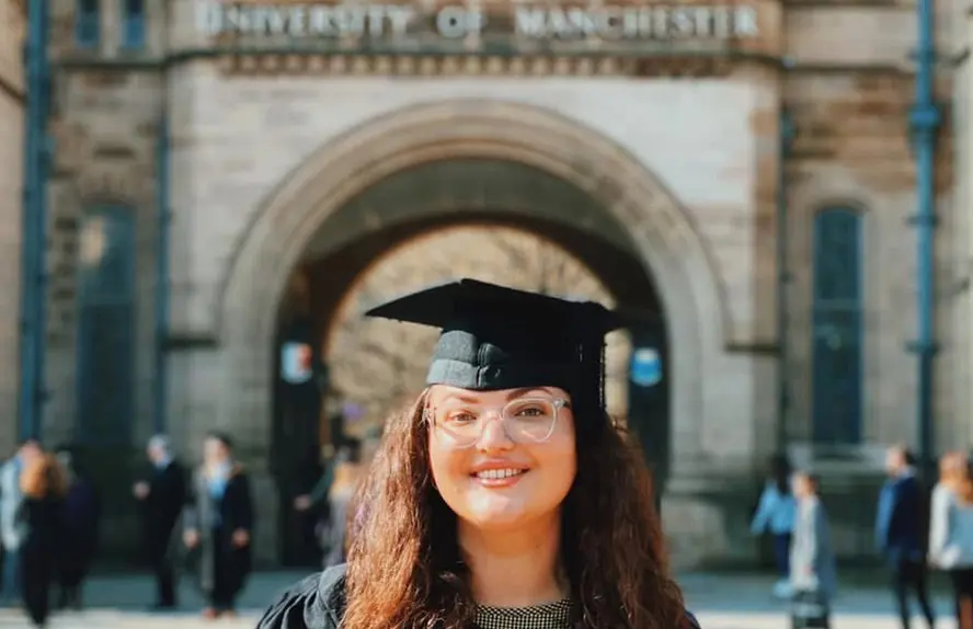 Graduate Becca Harrington wearing cap and gown standing in front of the University of Manchester entrance on graduation day.