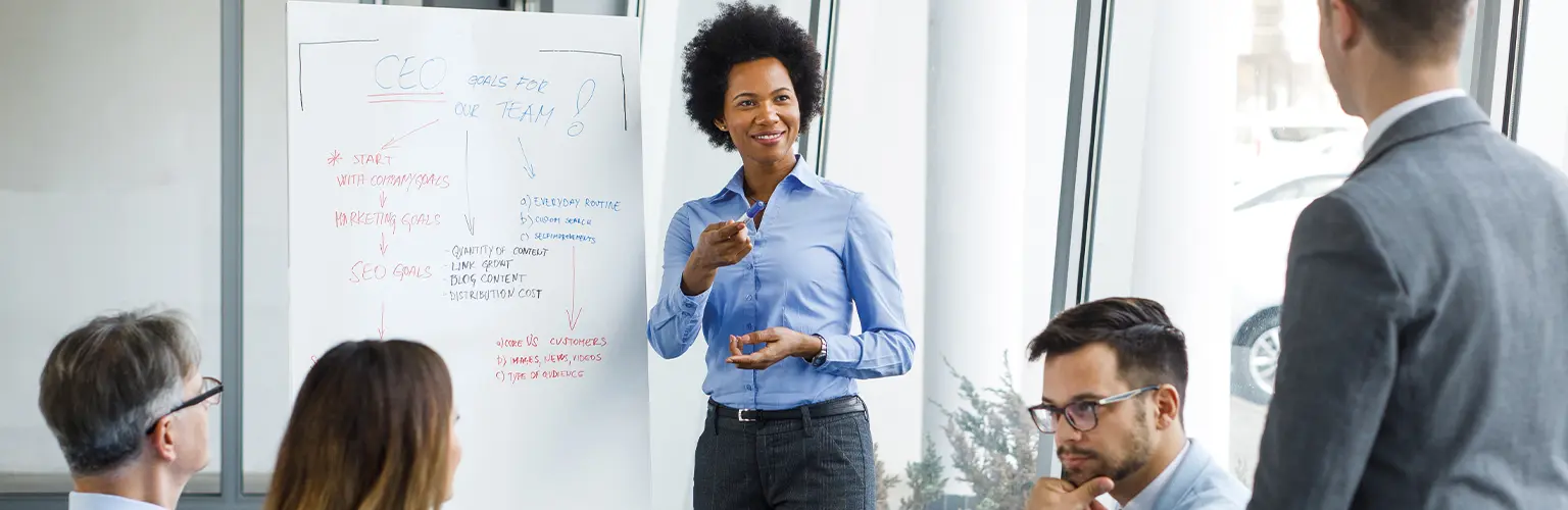 Woman leading on a meeting stood next to a white board and colleagues sat around a table