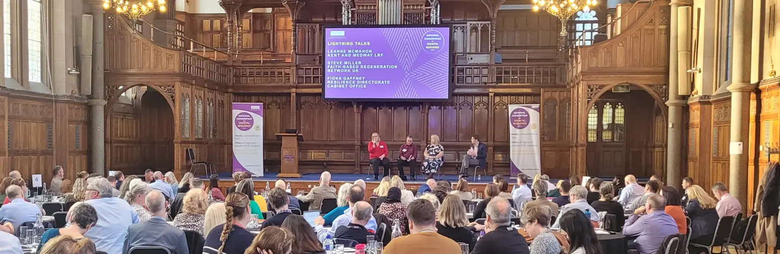 Large audience seated attending the National Conference on Societal Resilience. Four speakers sit on stage beneath a screen while chandeliers hang overhead and natural light streams through tall stained-glass windows.