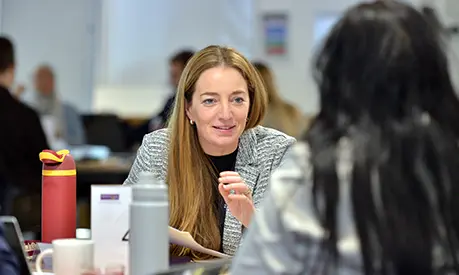 A female professional sat in a classroom talking to one of her peers across the table