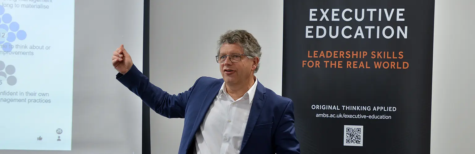 Professor Bart van Ark delivering a learning session at the front of a classroom next to a sign promoting Alliance Manchester Business School's executive education offering