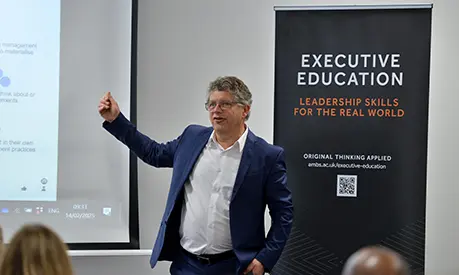 Professor Bart van Ark delivering a learning session at the front of a classroom next to a sign promoting Alliance Manchester Business School's executive education offering
