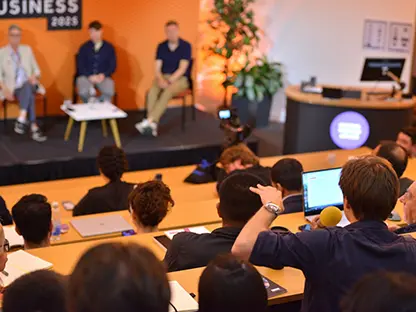 A large lecture room full of people attending a conference with three panellists on stage at the front of the room and a man in the audience asking a question.