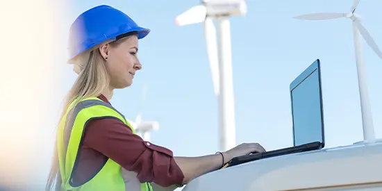 A woman with a clipboard wearing a hard hat and next to a wind turbine