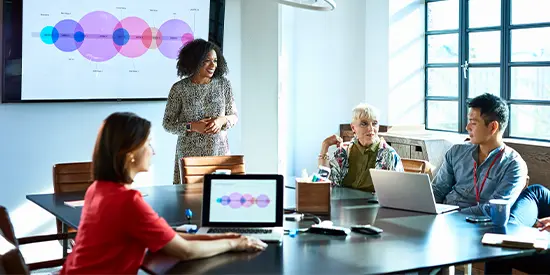 Colleagues sat around a table in a meeting with one person stood up in front of a screen at the front