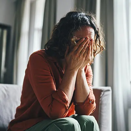 A model poses as a woman covering her face while sitting in a living room, conveying feelings of sadness or emotional stress.