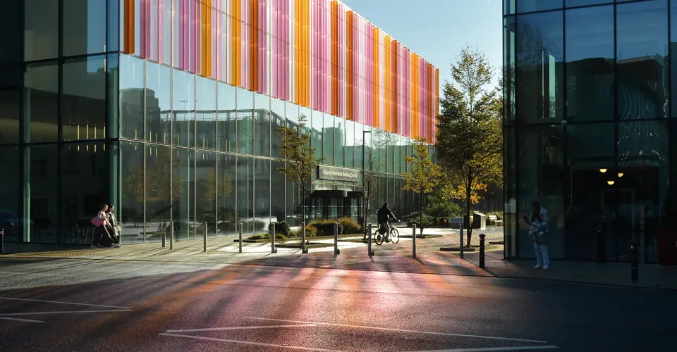 Exterior view of Alliance Manchester Business School with modern glass architecture and colorful vertical panels reflecting light onto the pavement.