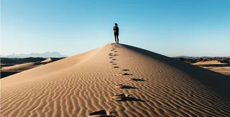 A woman walking up a hill in a desert, leaving foot steps behind her.