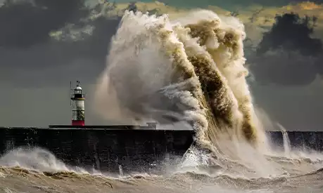 A large wave crashing into the side of a harbour near a lighthouse