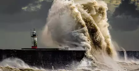 A large wave crashing into the side of a harbour near a lighthouse