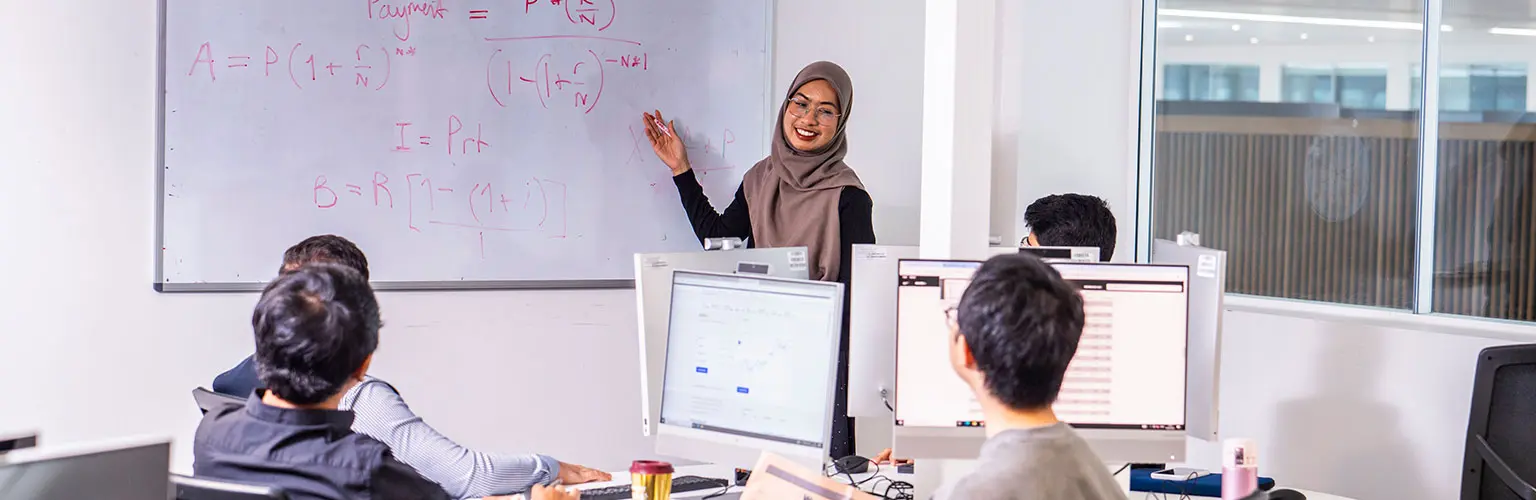 A student wearing a hijab presents mathematical formulas on a whiteboard to a group of classmates working on computers.