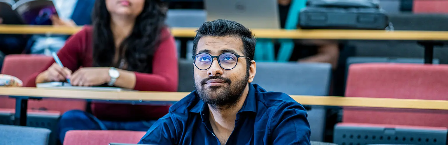 A student in glasses sits attentively in a lecture hall, listening and taking notes.