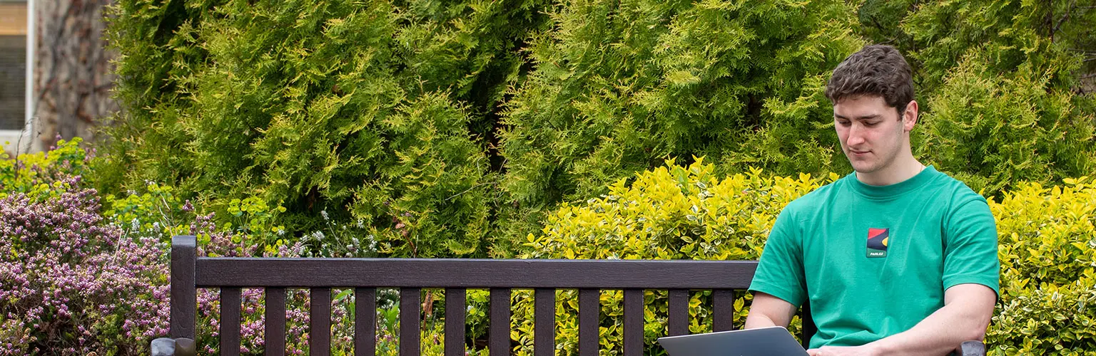 A student sits on a bench outdoors, working on a laptop surrounded by greenery.