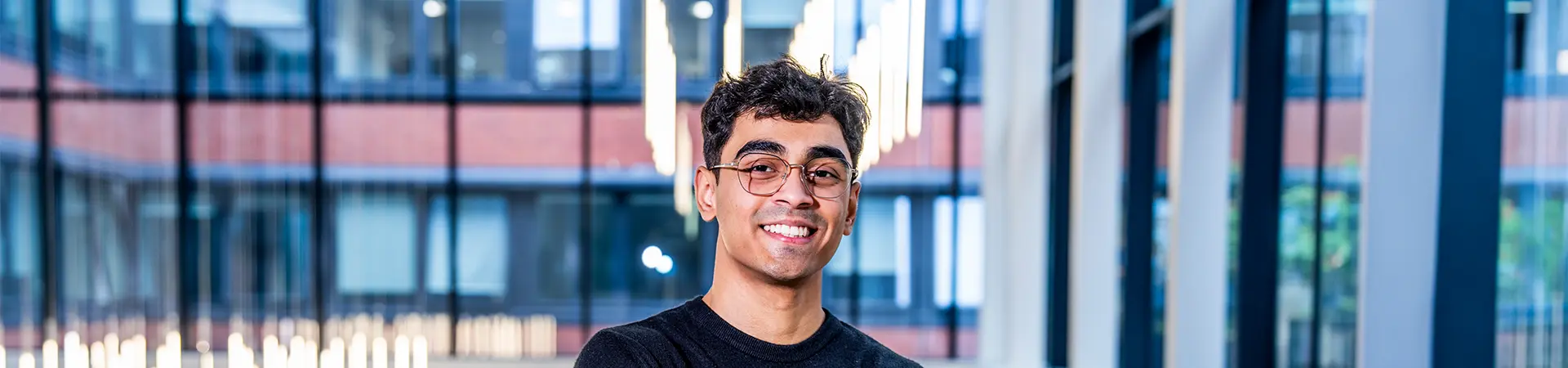 A student wearing glasses smiles while standing indoors at Alliance MBS.