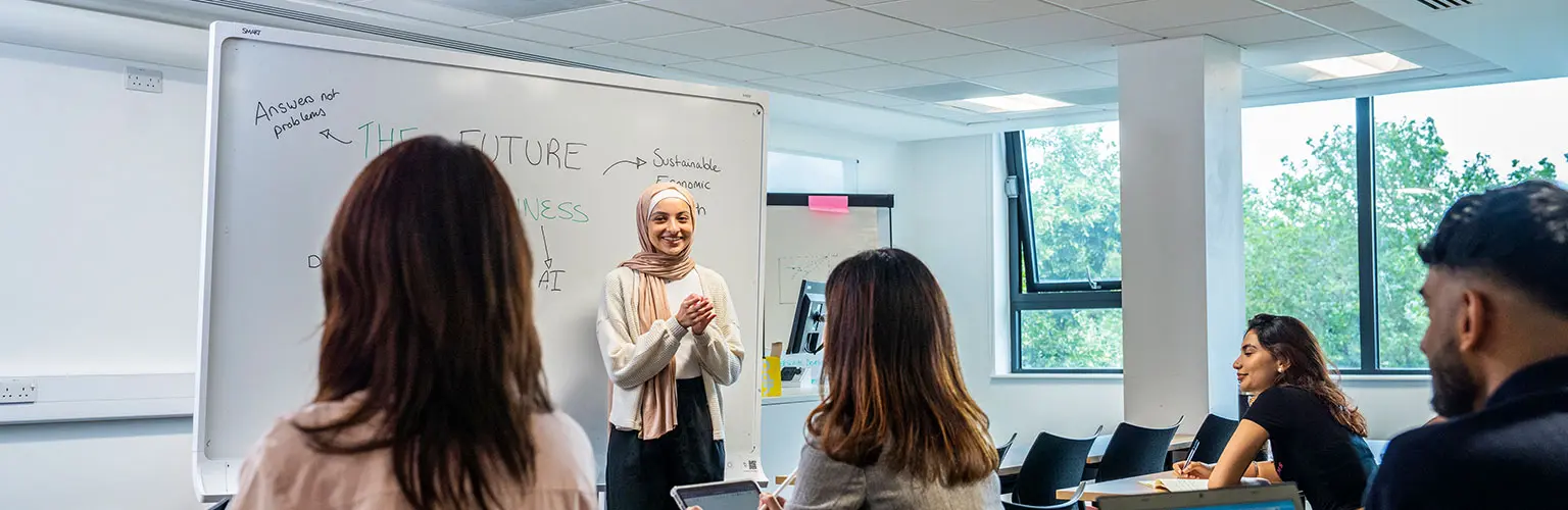 A student presents in front of a whiteboard while classmates listen and take notes.
