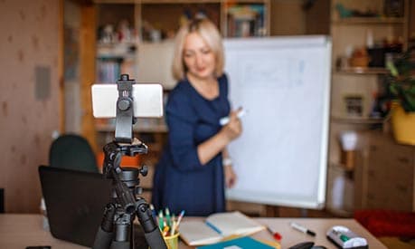 A woman teaching online using a physical whiteboard whilst a smartphone records