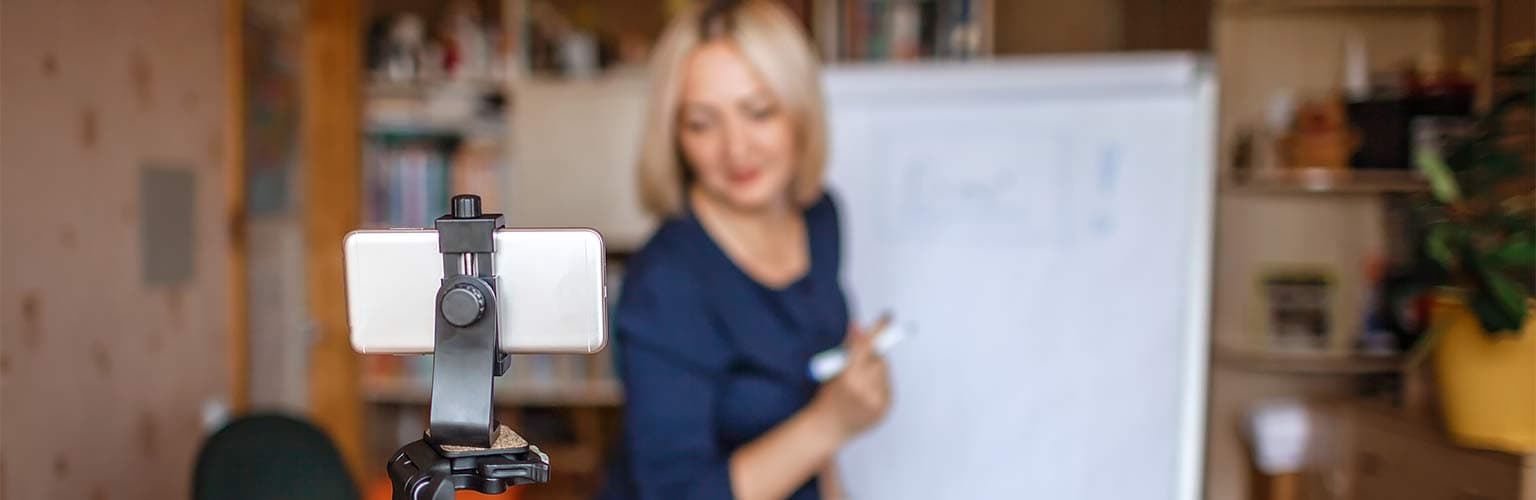 A woman teaching online using a physical whiteboard whilst a smartphone records