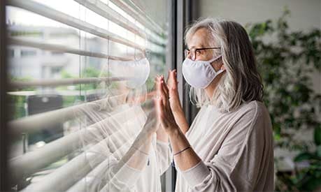 An elderly woman looking out of a window with a facemask on
