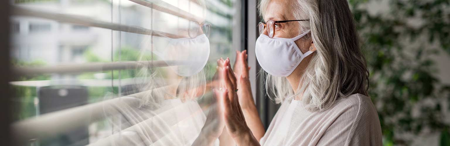 An elderly woman looking out of a window with a facemask on