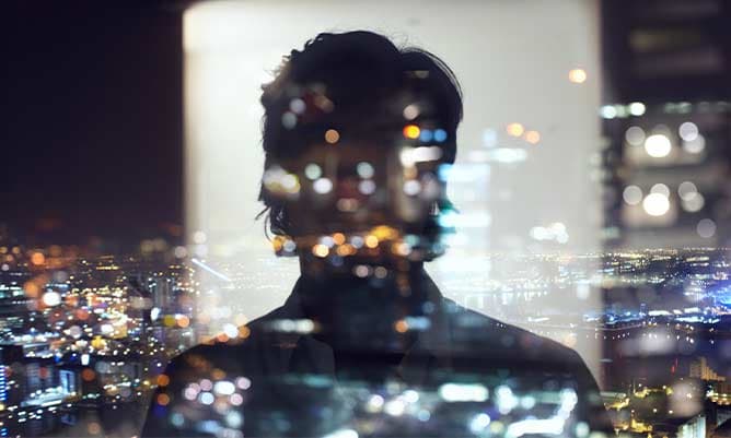 A silhouette of person reflecting on glass looking out over a cityscape at night