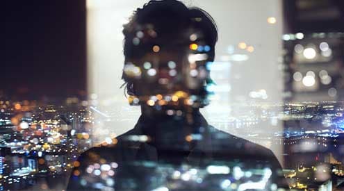 A silhouette of person reflecting on glass looking out over a cityscape at night