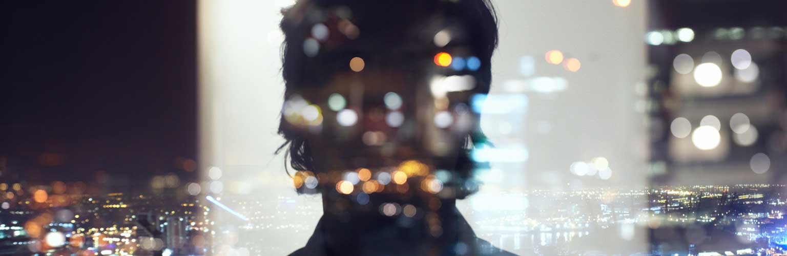 A silhouette of person reflecting on glass looking out over a cityscape at night