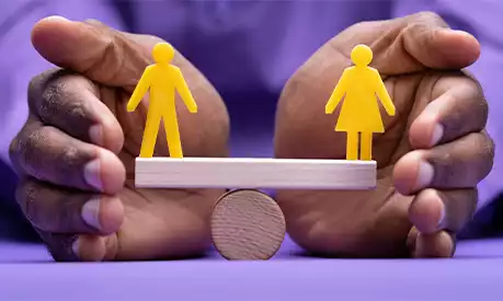 A pair of black women's hands balancing a piece of wood. On one side is a small yellow male figure and on the other is a small yellow woman figure. The wood is perfectly balanced, demonstrating them being equal to each other.