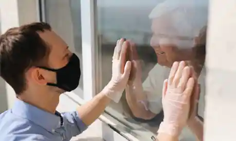 Man in face mask holds his hands up to the window of his Grandma, who holds her hands up to the glass too